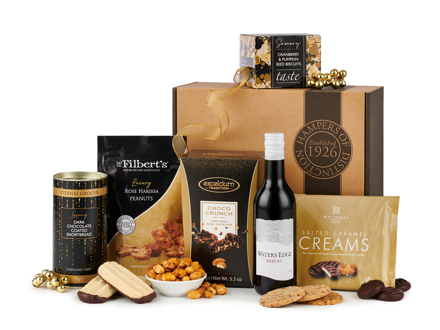 Red wine and treats gift box - spicers of hythe. With Merlot from Waters Edge, Salted Caramel Creams in golden packaging, chocolate dipped shortbread, rose harissa peanuts, pumpkin & cranberry seed biscuits sat in front of their Hampers of Distinction 1926 packaging.